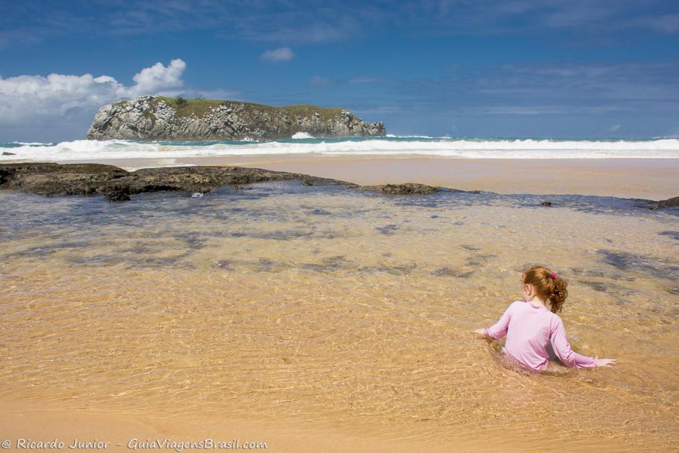 Imagem de uma criança sentada na beira do mar brincando.